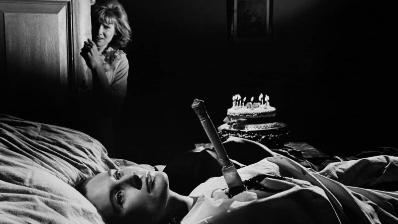 Imagem em preto e branco, uma mulher está deitada com uma faca enfiada em seu peito, ao seu lado tem um bolo de aniversário com as velas acesas e ao fundo há uma mulher olhando com expressão confusa. Darkflix