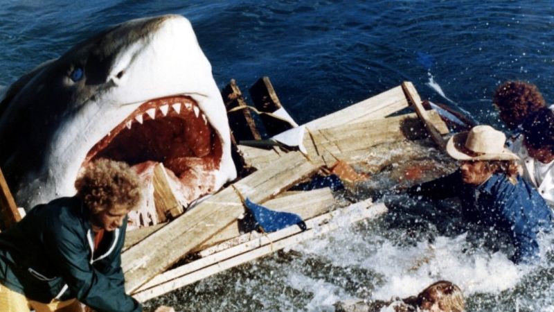 Um tubarão gigante destruindo algo de madeira, já indescritível, enquanto várias pessoas fogem.
